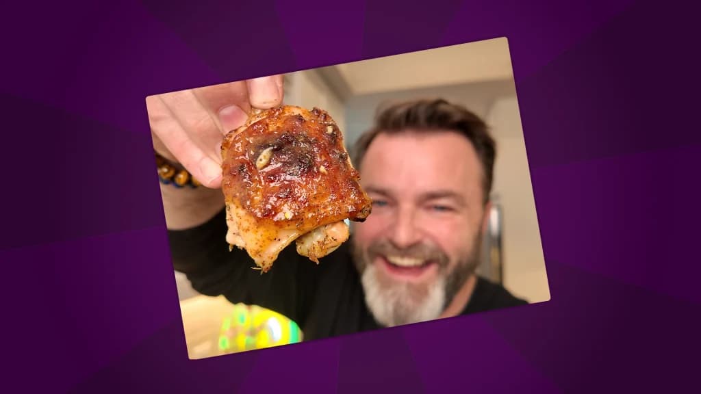 Close-up of a crispy roasted chicken thigh held up to the camera, with a smiling man blurred in the background.