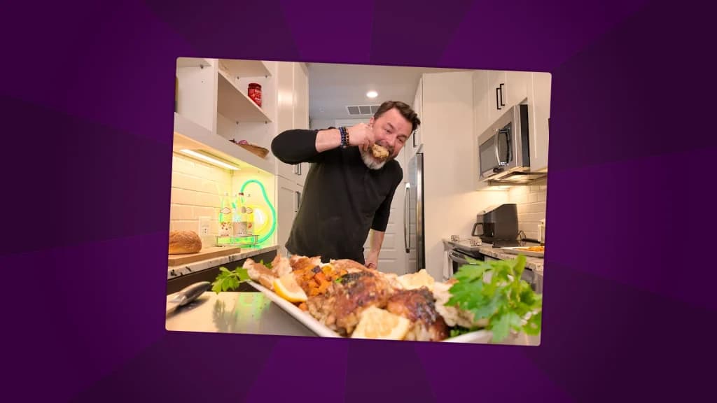 A man taking a bite of a roasted chicken in a bright home kitchen, with a large platter of chicken and vegetables in the foreground.