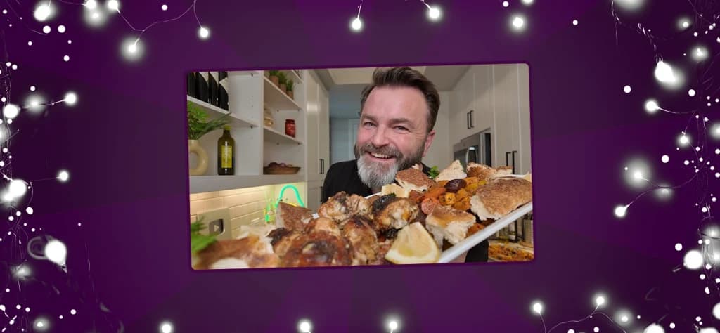 Smiling man holding a platter of roasted chicken and vegetables towards the camera in a modern kitchen.
