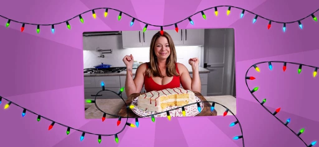 Woman smiling with her arms raised in celebration behind a large, sliced Christmas tree-shaped cake on a plate; holiday lights frame the image.