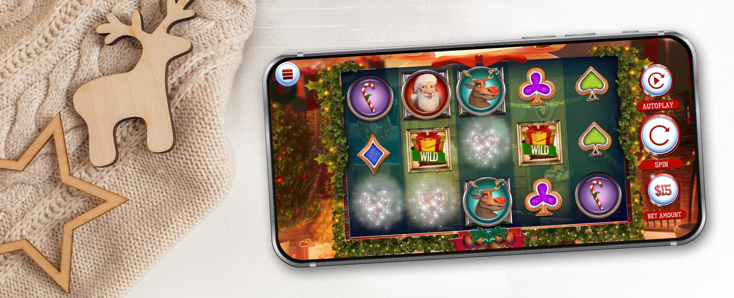 A mobile phone sits atop a white table, showing a screenshot of the Cafe Casino slot game “Santa’s Ways Hot Drop Jackpots” - showcasing the games reels and their features. To the left, is a beige knitted sweater with wooden cut-out shapes of a reindeer and a star.