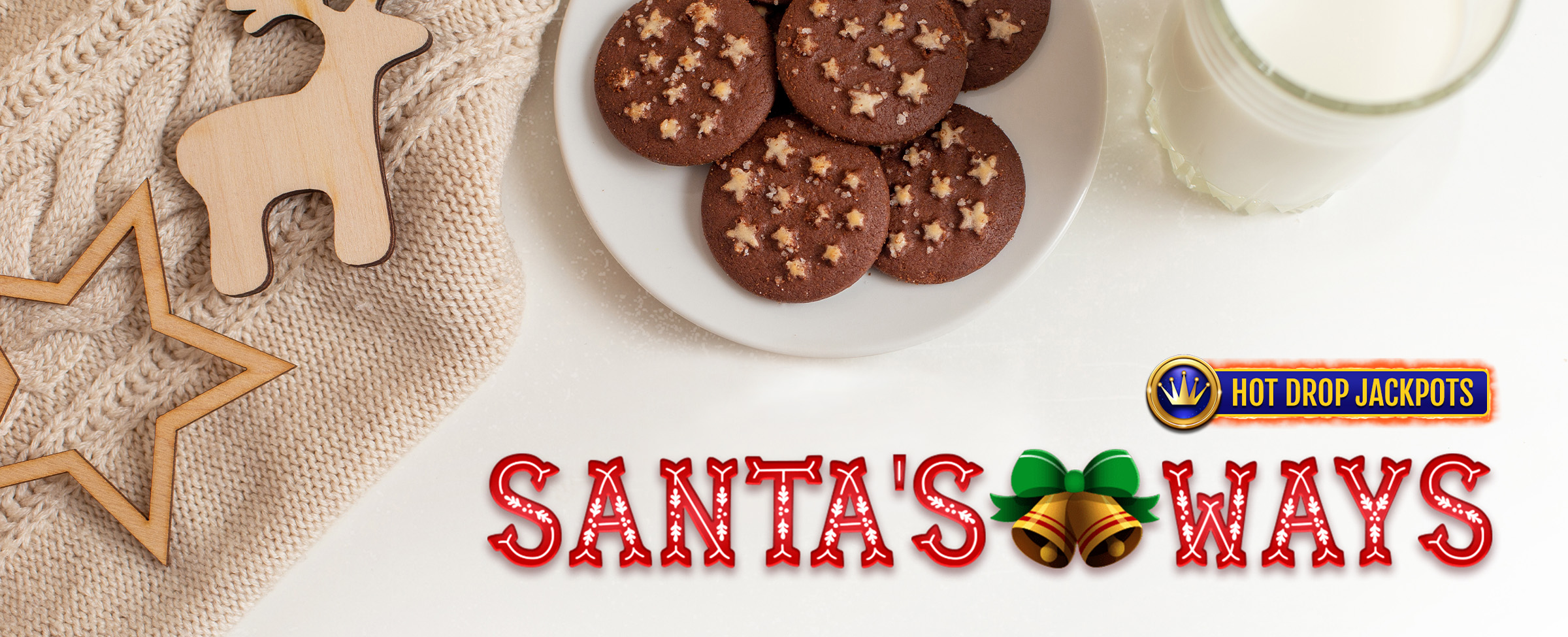 Chocolate cookies dusted in cocoa are piled on a white plate, sitting next to a white Christmas sweater with wooden cut-out shapes of a reindeer and a star, a glass of milk, and the Santa’s Ways Hot Drop Jackpots logo from the Cafe Casino slot game of the same name.
