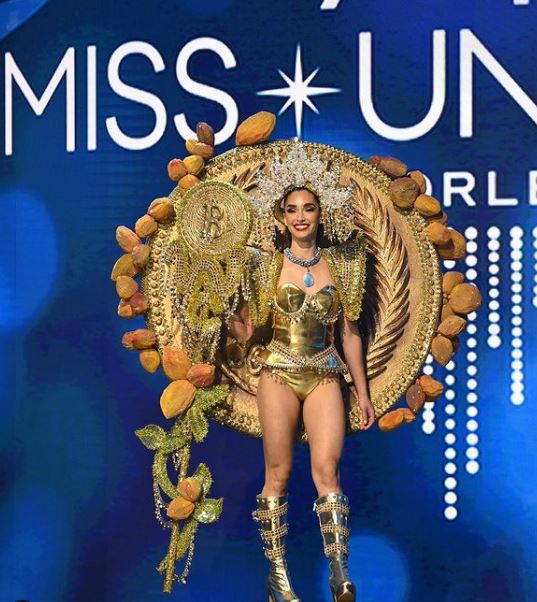Miss Universe El Salvador walking across the stage in her golden outfit framed by a golden coin on her back and a large golden staff with a Bitcoin at the top at her side.