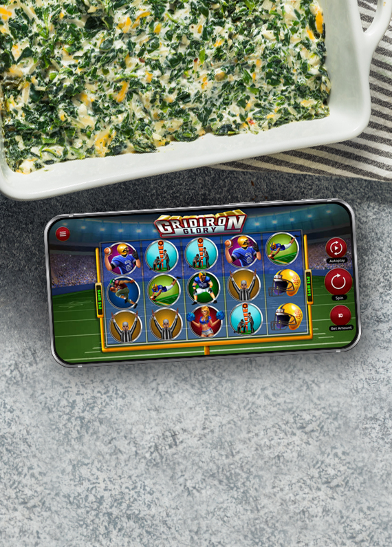 A mobile phone showing a screen from Cafe Casino’s slot game, Gridiron Glory, sits on a table next to a baking dish filled with spinach dip.