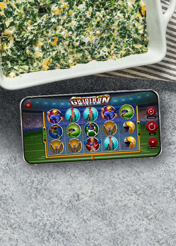 A mobile phone showing a screen from Cafe Casino’s slot game, Gridiron Glory, sits on a table next to a baking dish filled with spinach dip.