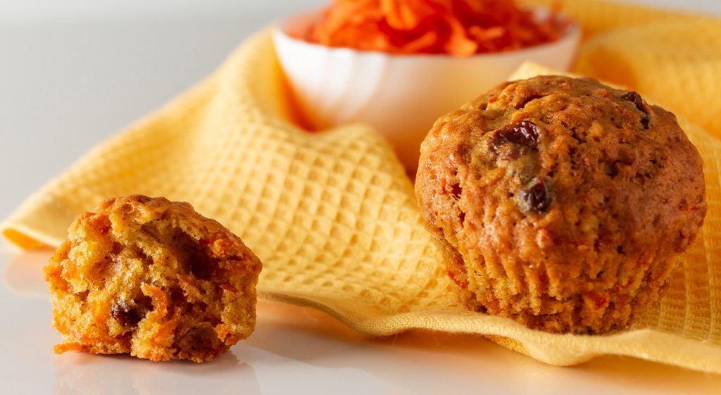 On top of a white high-gloss table surface sits a yellow tea towel, with a small white bowl filled with grated carrot, and in front, two carrot muffins.