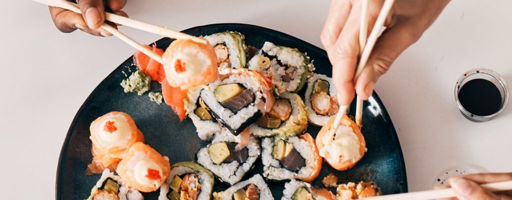 A large black share platter is seen on a white bench surface, hosting a selection of sushi rolls and sashimi, with three hands using chopsticks to pick up their selection.