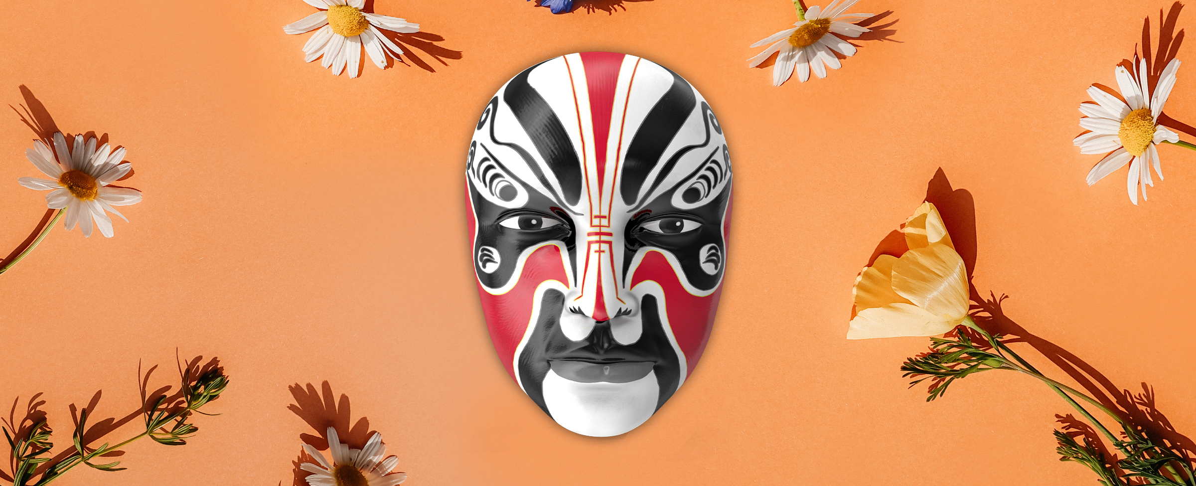 A red, black, and white mask is seen, from the Cafe Casino online slot Opera of the Mask, surrounded by various flowers which are resting on top of an apricot-colored table surface.