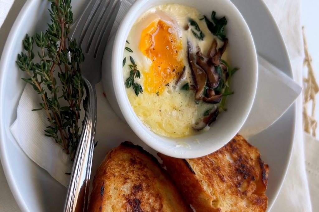 Eggs en Cocotte or baked eggs sit in a white bowl, on top of a white plate featuring a crusty bread and a fork.