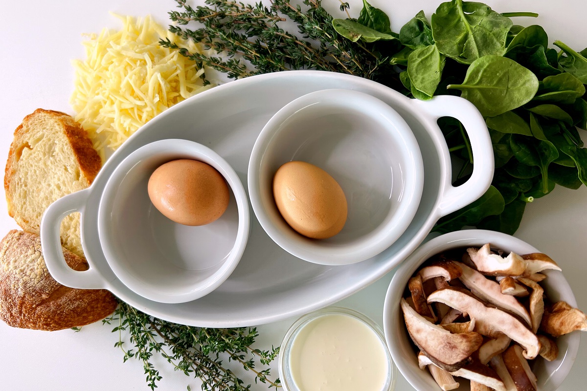 Ingredients for this Cafe Casino sponsored recipe, sit on a white counter: spinach, shredded cheese, thyme, bread, mushrooms in a small dish, and un-cracked eggs in a small dish.