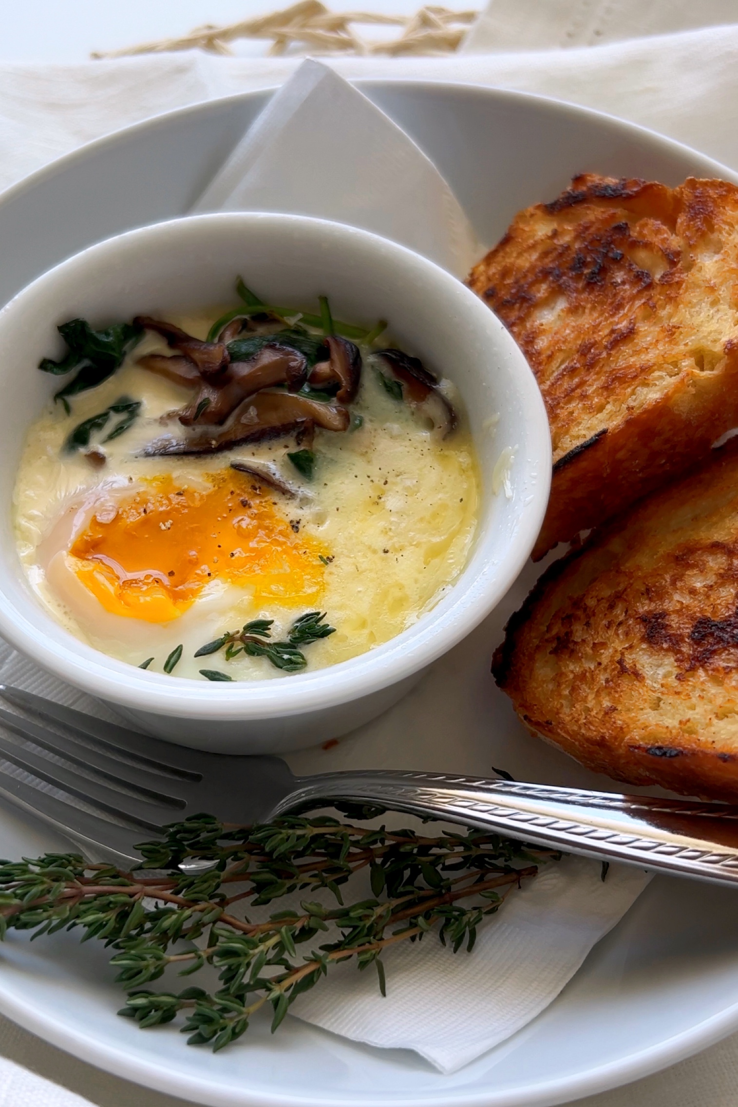 Eggs en Cocotte or baked eggs sit in a white bowl, on top of a white plate featuring a crusty bread and a fork.