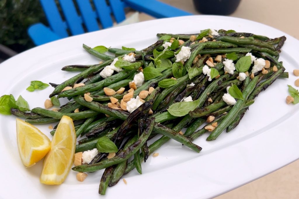 The raw ingredients for The Hunger Diaries’ summer recipe sit on a white, square plate adjacent to a barbecue grill: green beans in a glass bowl, basil, almonds, lemon halves and goat cheese crumble.
