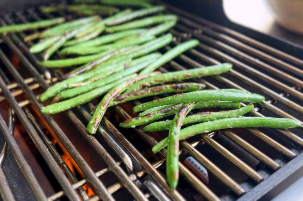 Lightly salted and oiled green beans laid across a barbecue grill.