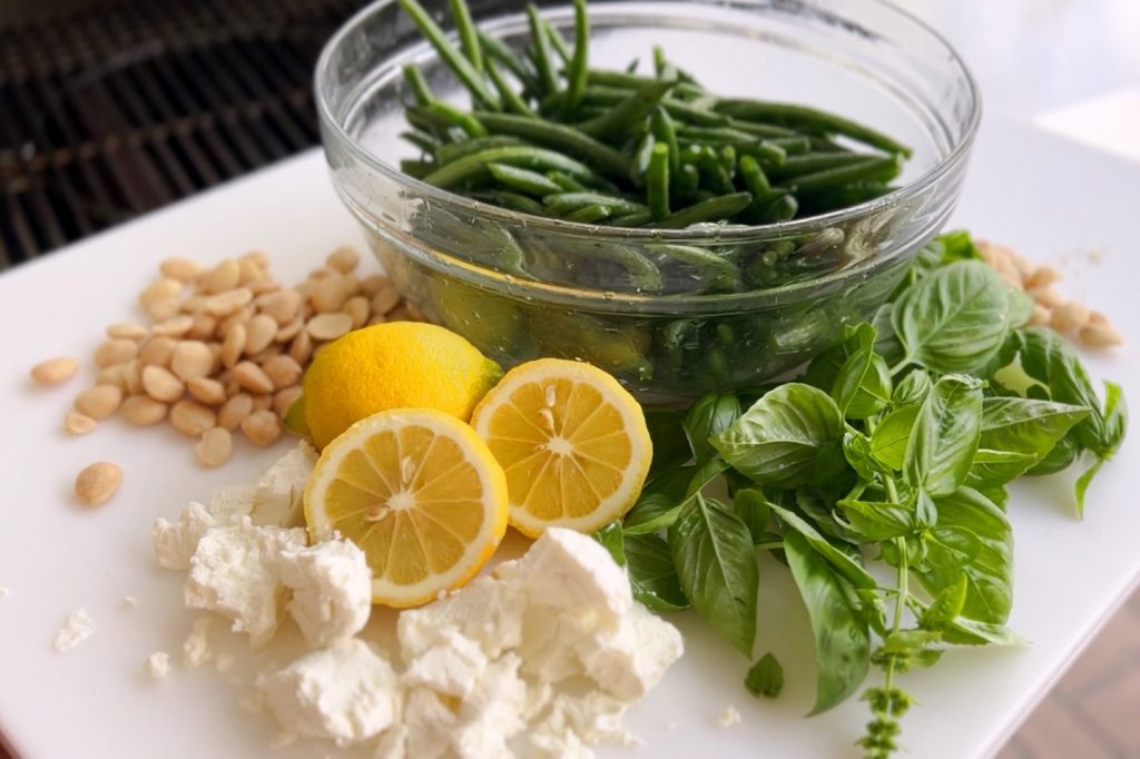 The raw ingredients for The Hunger Diaries’ summer recipe sit on a white, square plate adjacent to a barbecue grill: green beans in a glass bowl, basil, almonds, lemon halves and goat cheese crumble.