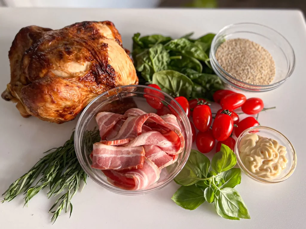 Ingredients for this Cafe Casino sponsored recipe, sit on a white counter: a roasted whole chicken, spinach leaves, quinoa in a small glass bowl, uncooked bacon in a larger glass bowl, a handful of cherry tomatoes, mayo in a small glass bowl, and assorted herbs.