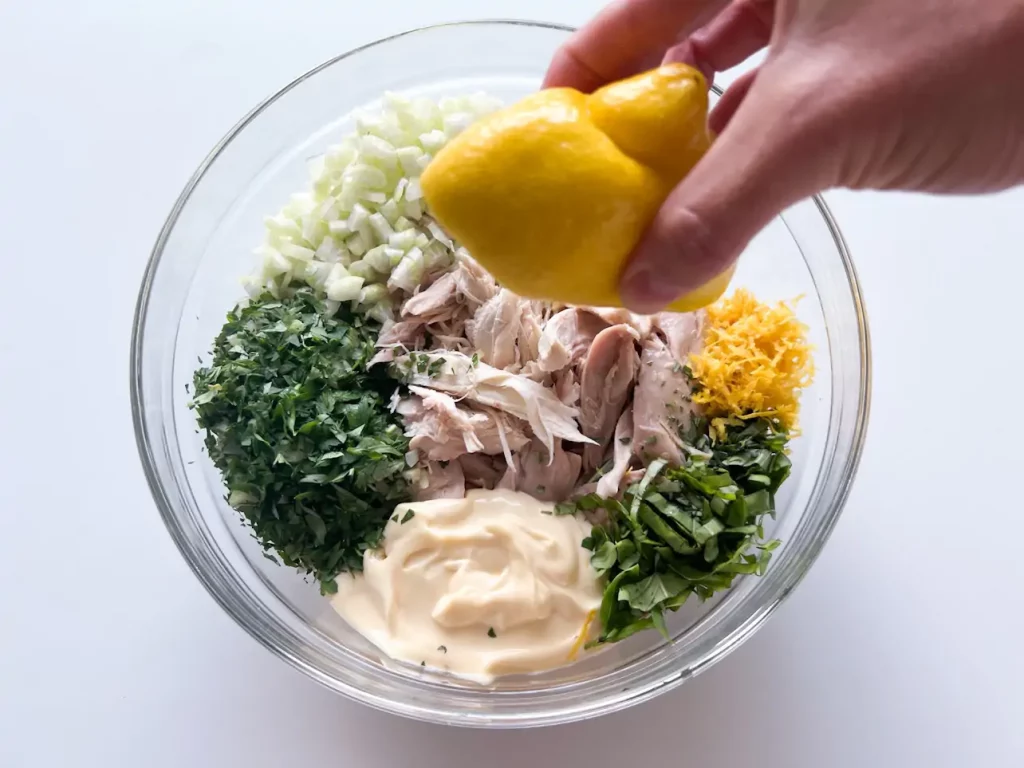 An image featuring Lindsay Moser’s hand squeezing a lemon over a glass dish featuring shredded chicken, mayo, diced spinach, and other ingredients; based on the recipe for Cafe Casino.