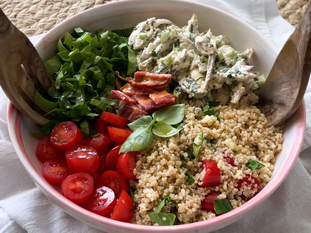 A large serving bowl featuring ingredients based on the recipe for Cafe Casino by Lindsay Moser; cherry tomatoes, diced spinach, crispy bacon pieces, chicken salad, and quinoa (clockwise from bottom left). A large wooden salad fork and spoon are on either side of the bowl.