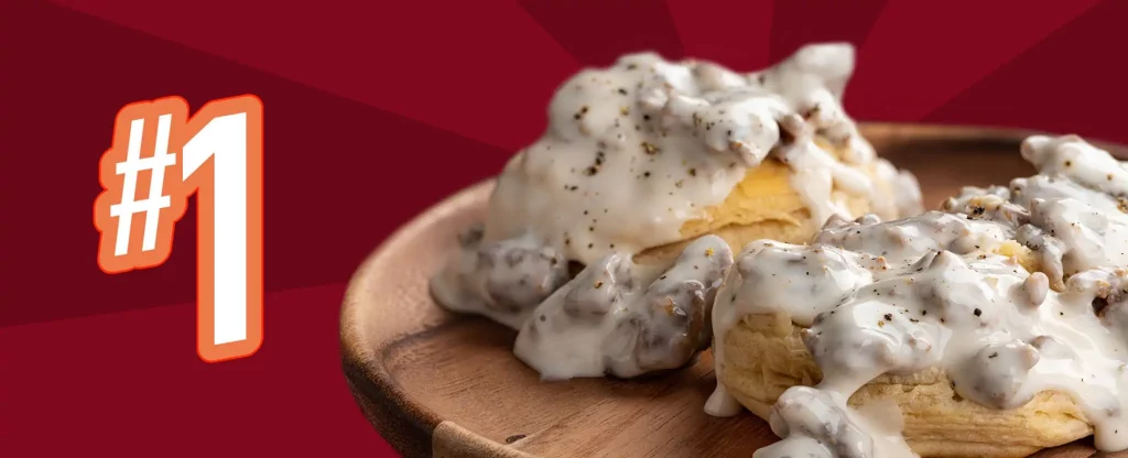We see a dish of traditional biscuits and gravy sitting in a shallow wooden bowl. The thick, corned biscuits are topped with creamy sausage gravy. To the left we see a “#1”, in large, bold font, with a thick orange border. Behind, is a background in hues of maroon and dark red.