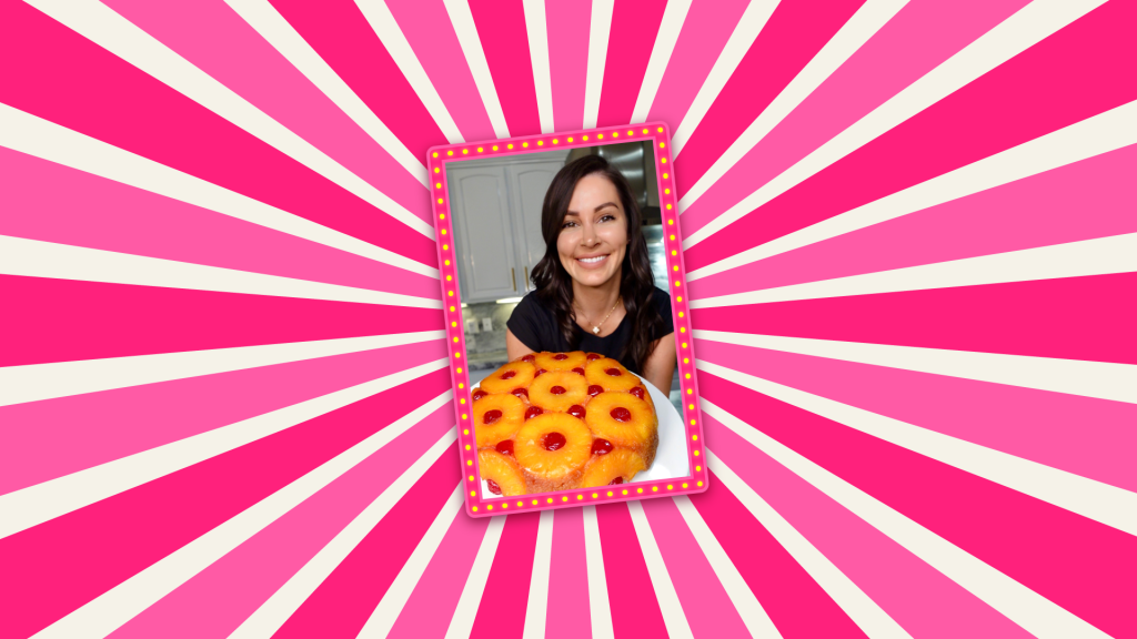 Chef Genevieve is wearing a black top, holding a white dish with a Pineapple Upside-Down Cake presented on top; set on a pink striped background.