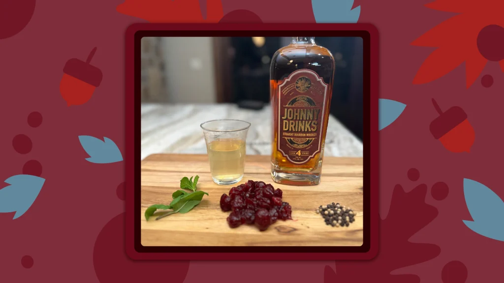 A Johnny Drinks bottle of bourbon on a wooden cutting board with a glass of liquor, cranberries and mint leaves.