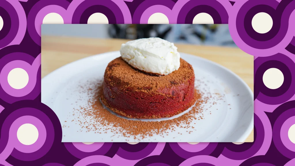 A round, red velvet lava cake, with a dollop of cream on top and a dusting of dark chocolate cocoa powder on a white plate.