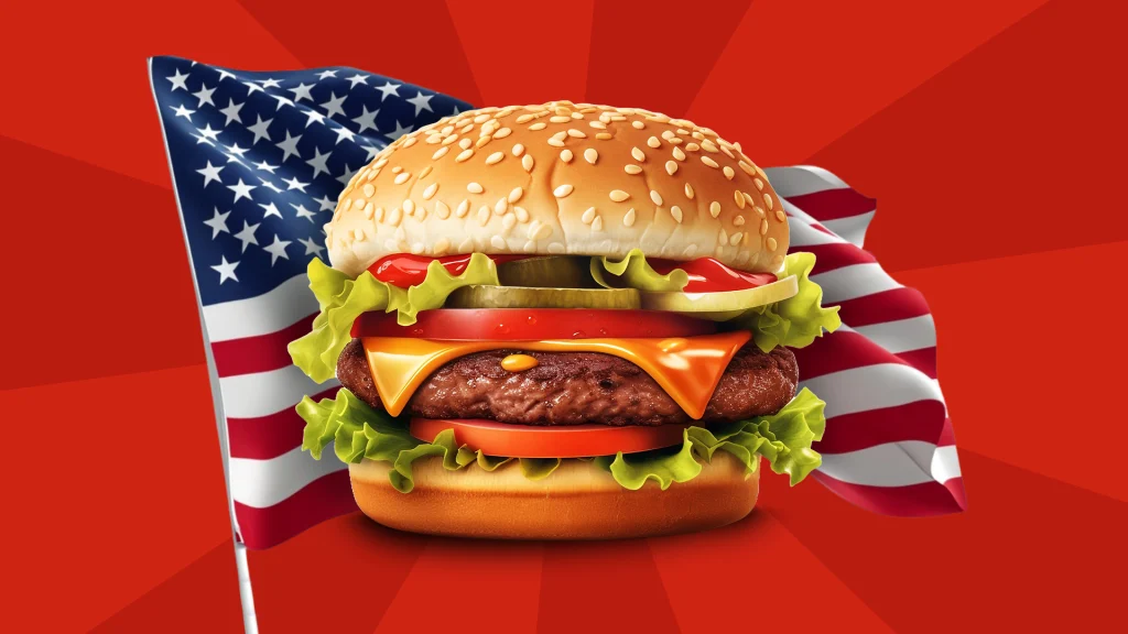 A huge burger is in front of an American flag and a red backdrop.