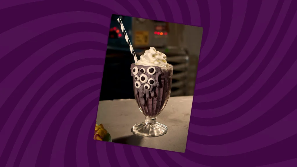 a blueberyy milkshake topped with whipped cream sits on a ourple background. The glass is flowing over with melted purple chocolate and candy eyeballs sticking out