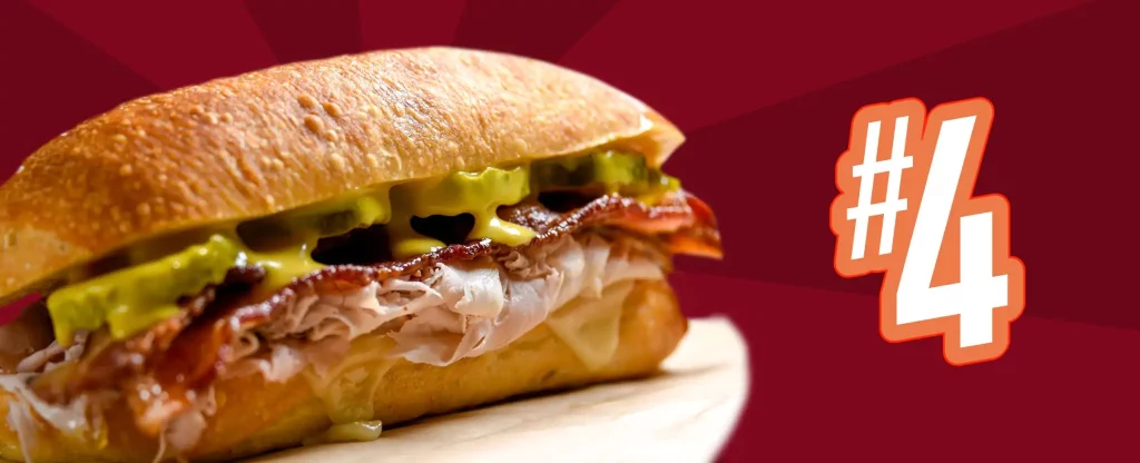 To the left of the image is a Reuben Sandwich, made up of a long roll, slices of meat, pickles, drenched in dressing. To the right we see a “#4”, in white, bold type and a thick orange border. In the distance is a red and maroon-toned background.